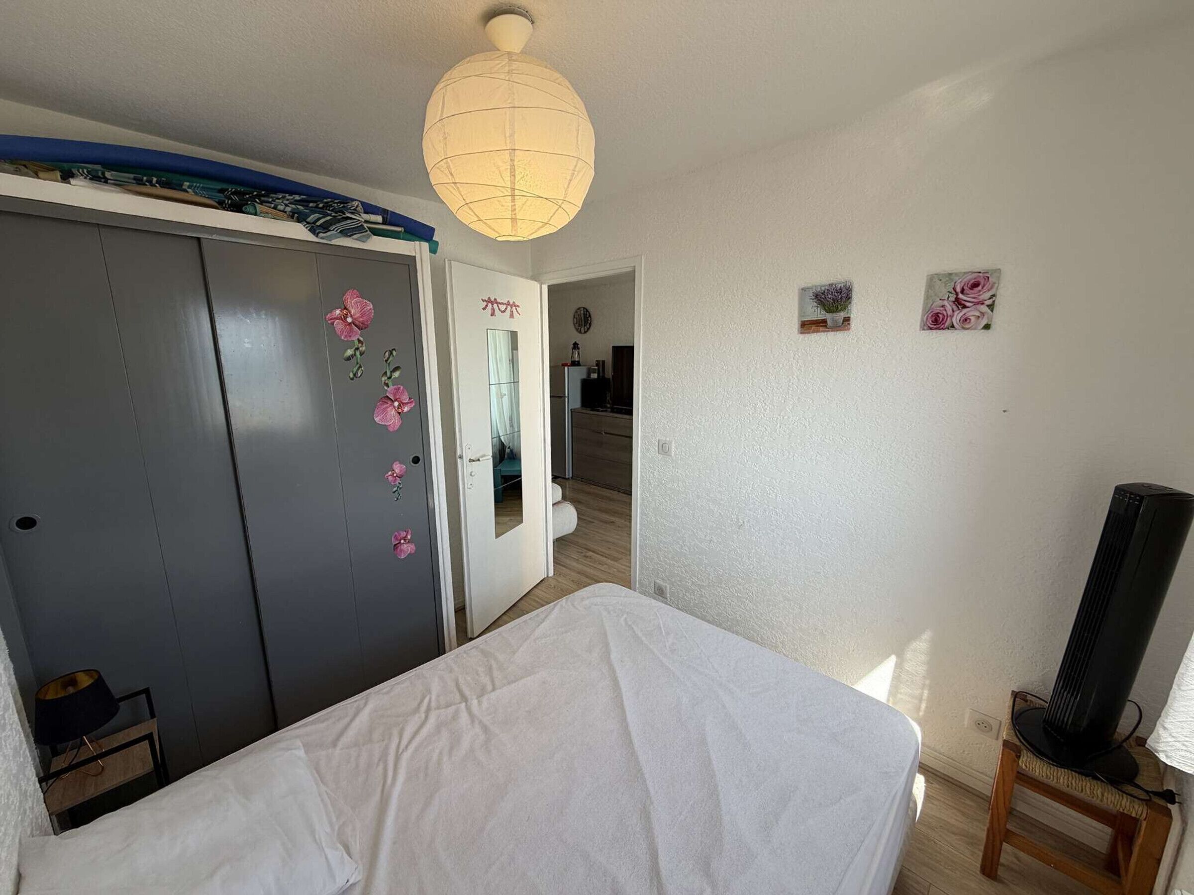 A bedroom with a bed, a wardrobe with floral stickers, a mirror, a ceiling light, and a small plant on the wall.