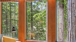 A room with large windows, a wooden wall, a lamp, and a sofa. - Moonraker House | Redwoods, Hot Tub & Trails (Sea Ranch)