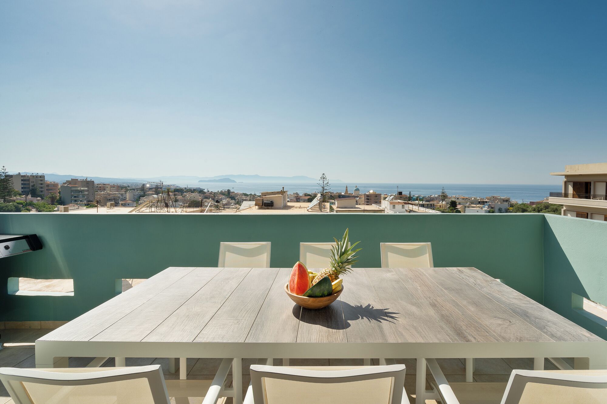 A rooftop terrace with a table and chairs, a bowl of fruit, and a view of the city and sea.