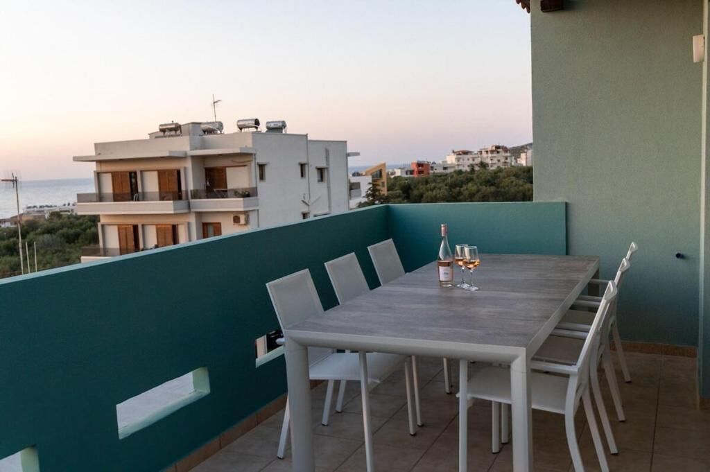 A balcony with a dining table set for four, white chairs, and a view of the sea and buildings.