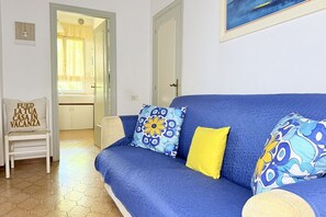 A blue sofa with patterned cushions, a white chair with a cushion, and a door leading to a bathroom. - M436 - Marcelli, three-room apartment with garden, 150 meters from the sea (Marcelli)