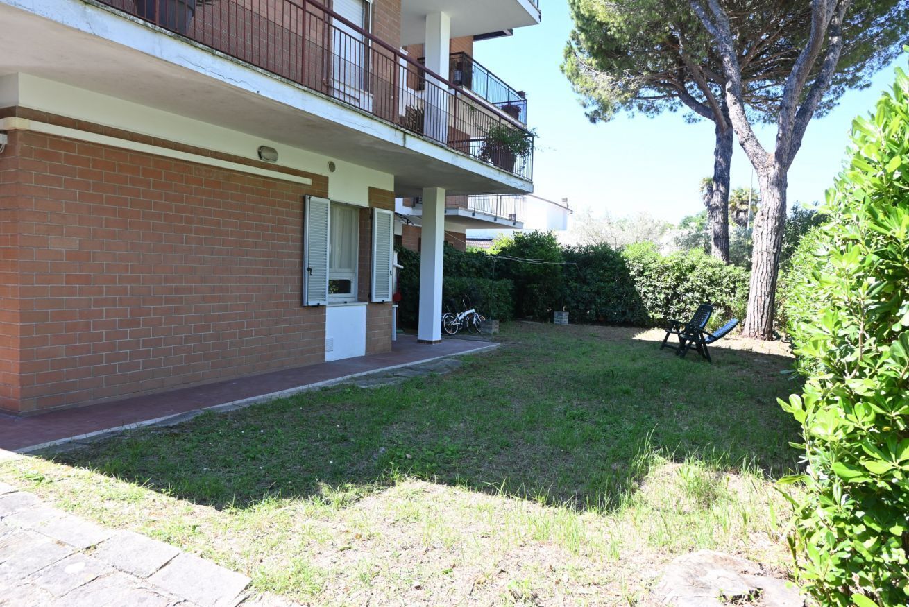 A residential building with a brick facade, a balcony with a metal railing, and a grassy yard with a tree and a chair.