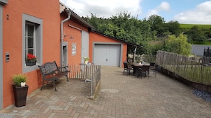 A building with a garage, a patio with a table and chairs, a bench, and a potted plant. - Eifel Dream Country House with Private Outdoor Hot Tub and Sauna (Plütscheid)