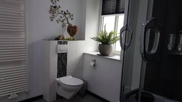 Un bagno con il water, una doccia, un termosifone, una finestra con una pianta e un elemento decorativo a parete.