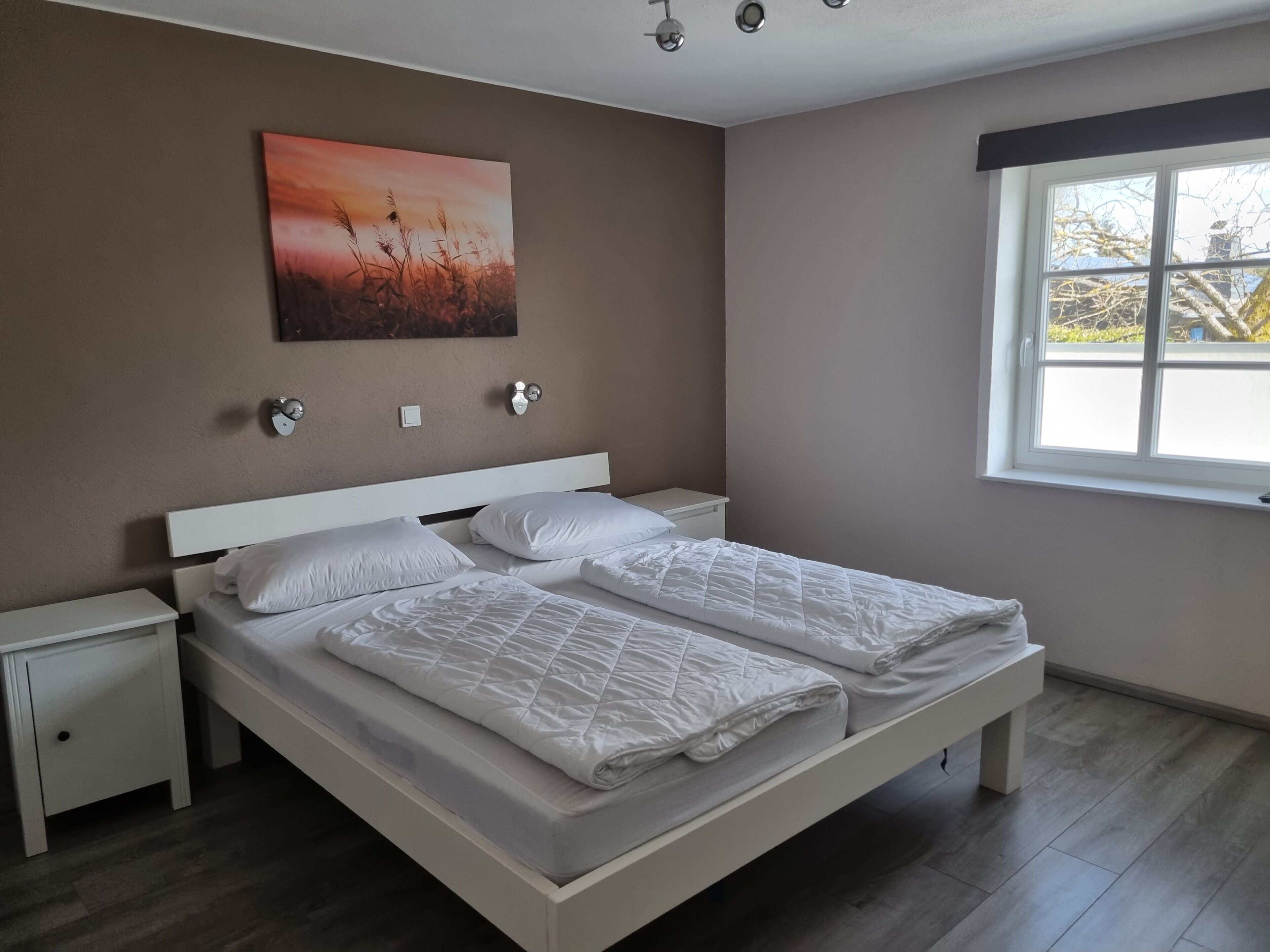 Un lit double avec une literie blanche, une table de chevet, une lampe fixée au mur et une fenêtre donnant sur des arbres.
