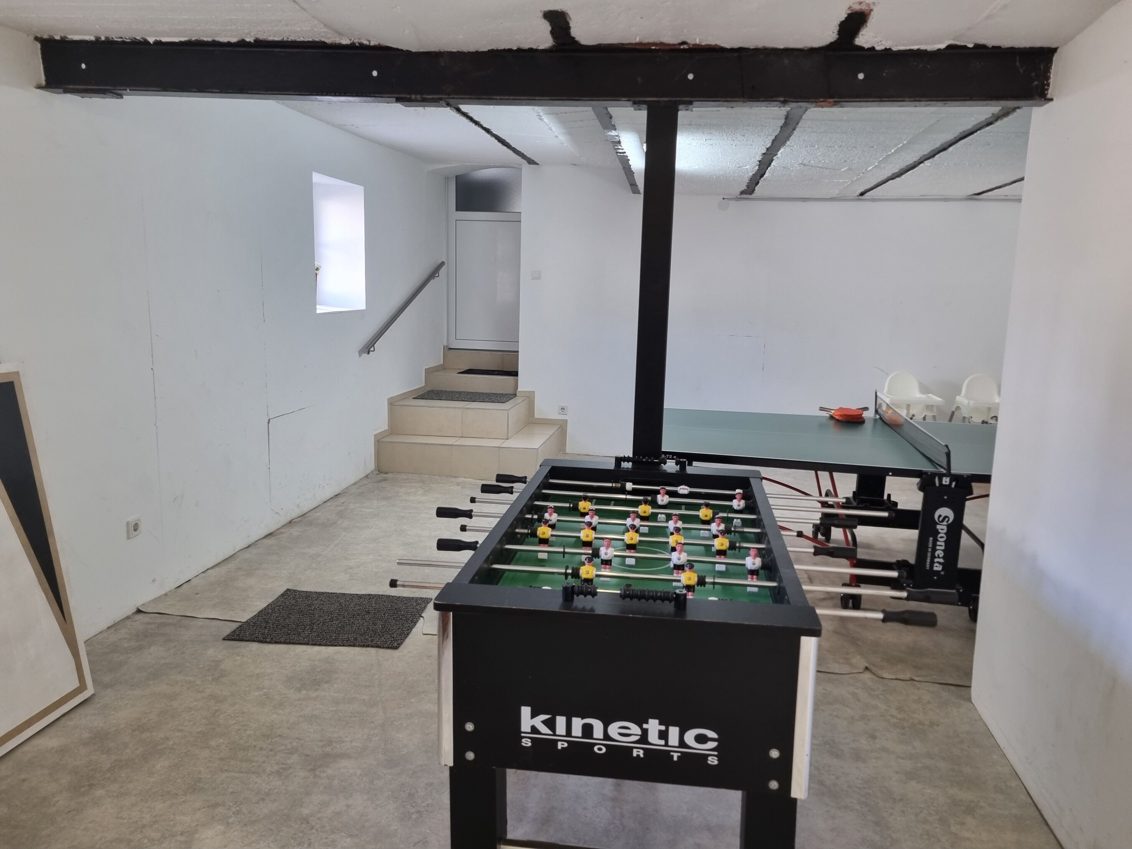 A room with a foosball table, a table tennis table, and a staircase.