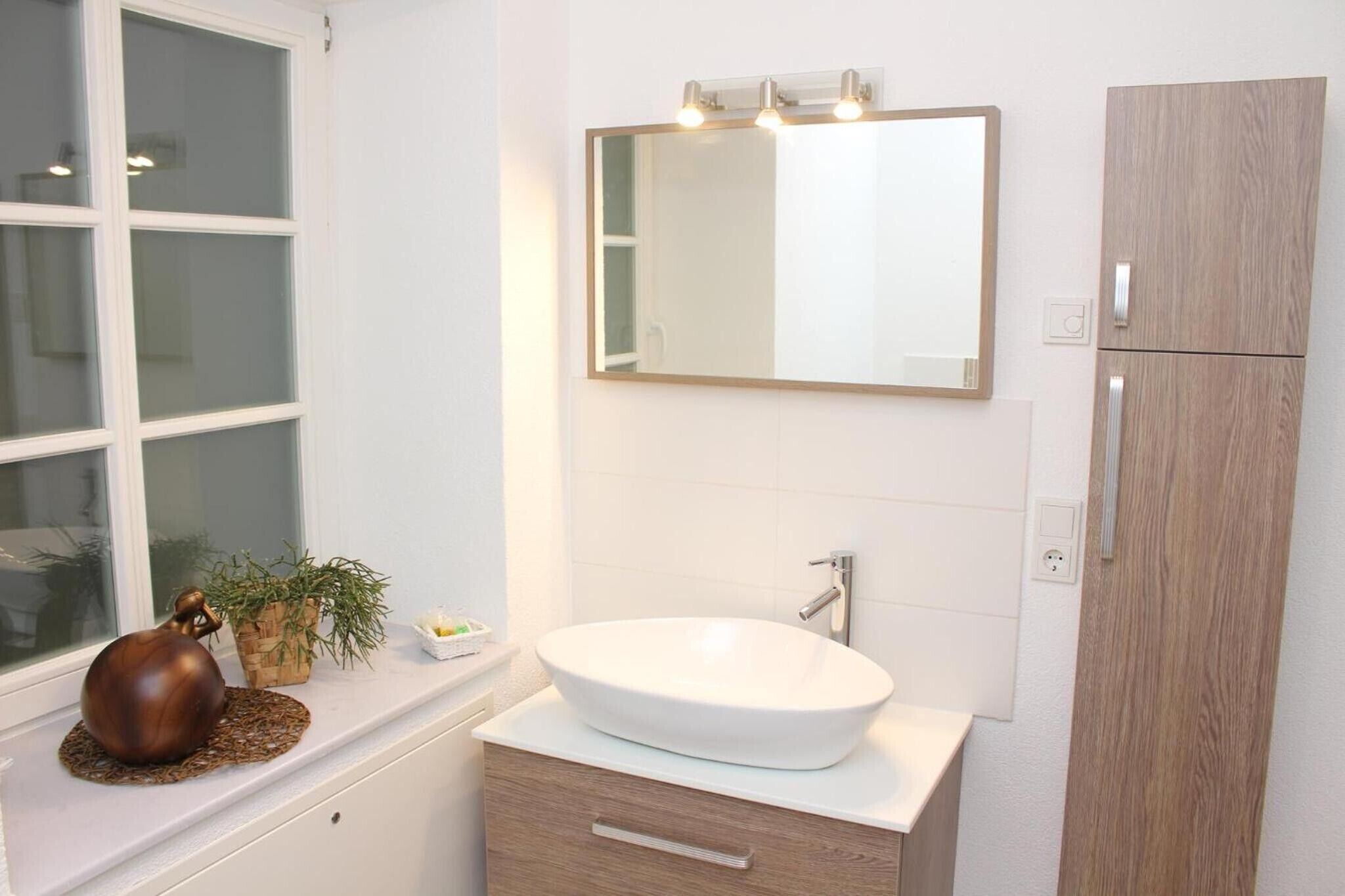 Ein Badezimmer mit einem weißen Waschbecken, einem beleuchteten Spiegel, einem Holzschrank und einem Fenster mit Jalousien.