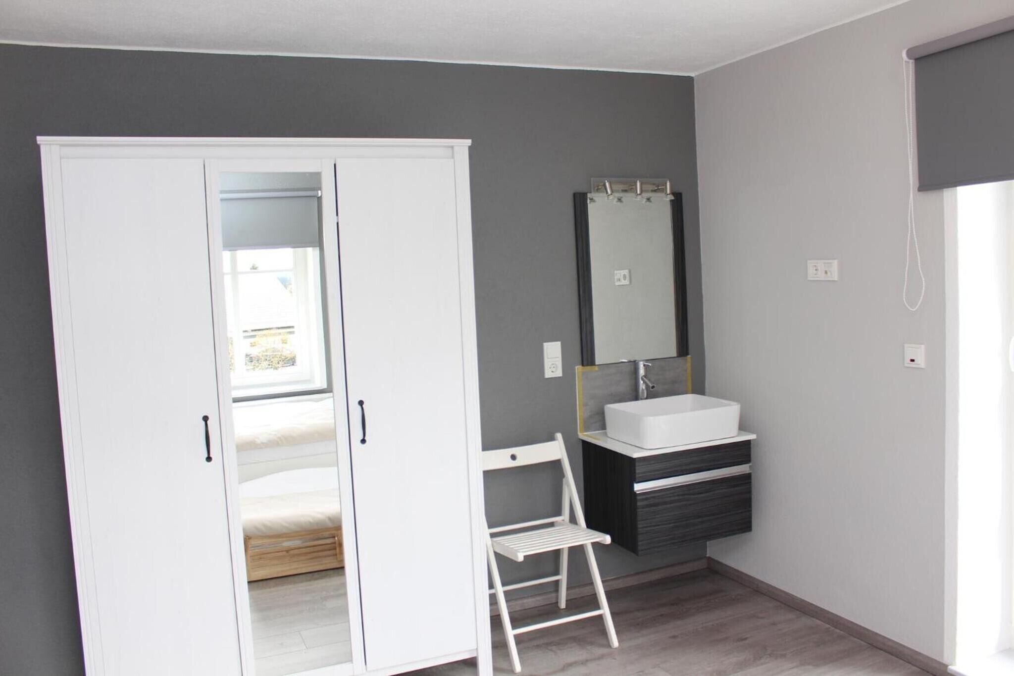 A bedroom with a wardrobe, a bed, a small white chair, a bathroom with a sink and mirror, and a window.