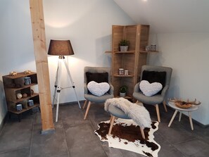 Ein gemütliches Zimmer mit einer Dreibein-Stehlampe, zwei grauen Sesseln mit herzförmigen Kissen, einer Holzbibliothek und einem kleinen runden Tisch mit einer Hirschdekoration.