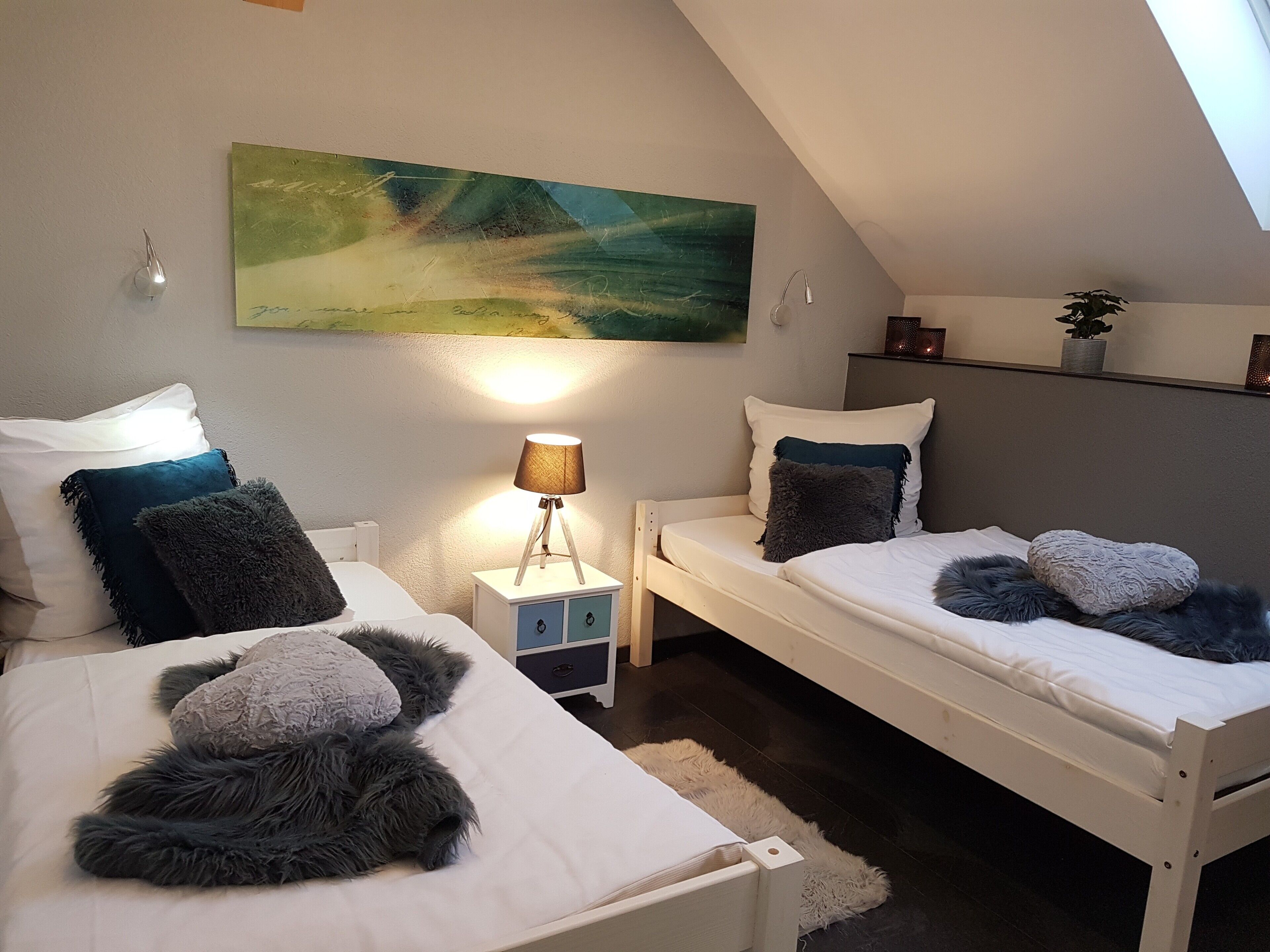 Un dormitorio con dos camas, una mesita de noche con lámpara, un cuadro en la pared y una planta en un estante.