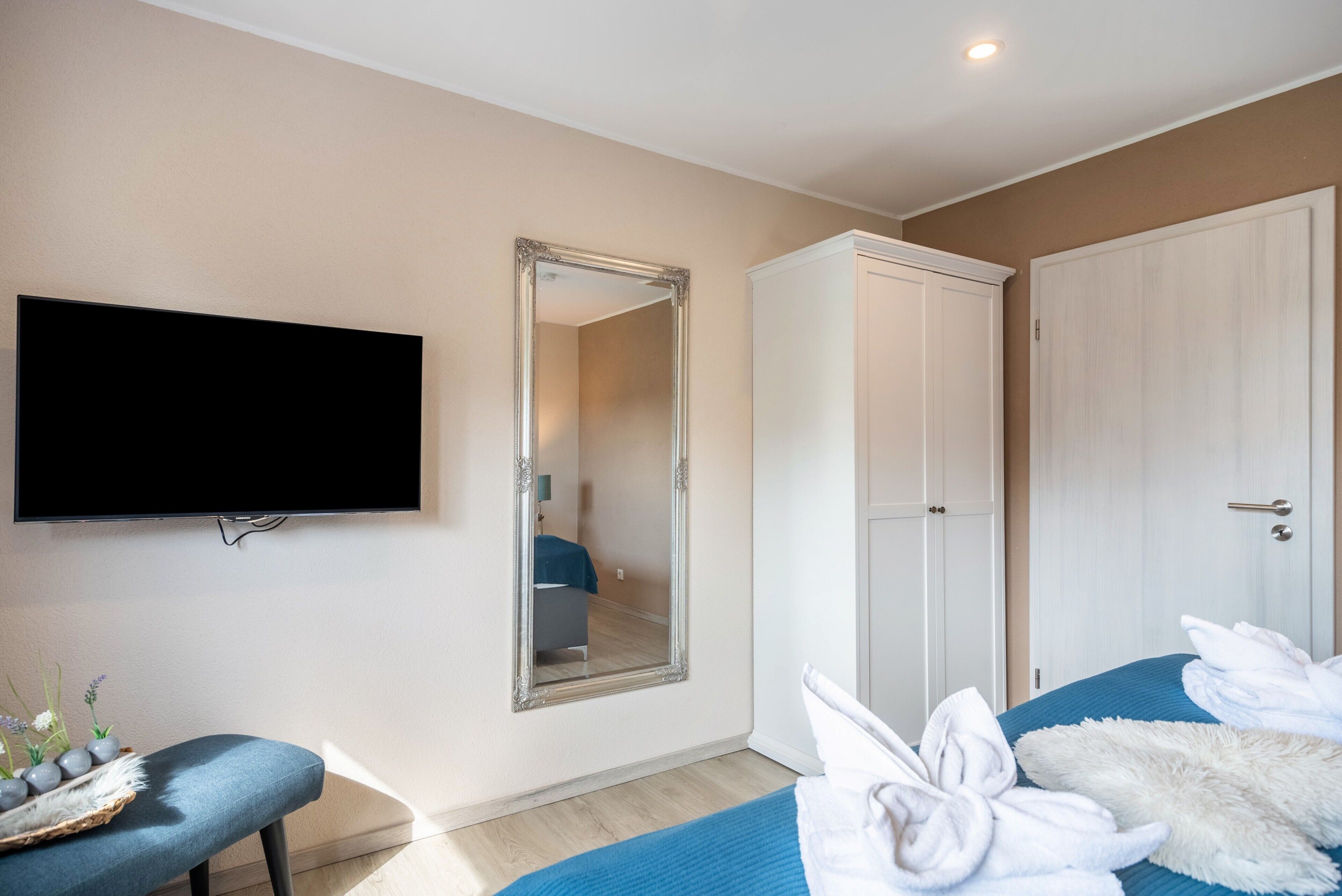 A hotel room with a flat-screen TV, a large mirror, a wardrobe, a bed with towels, and a bench with decorative items.