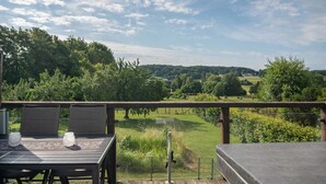 Une terrasse en bois avec une table et des chaises, donnant sur un paysage verdoyant, avec des arbres et une colline au loin.