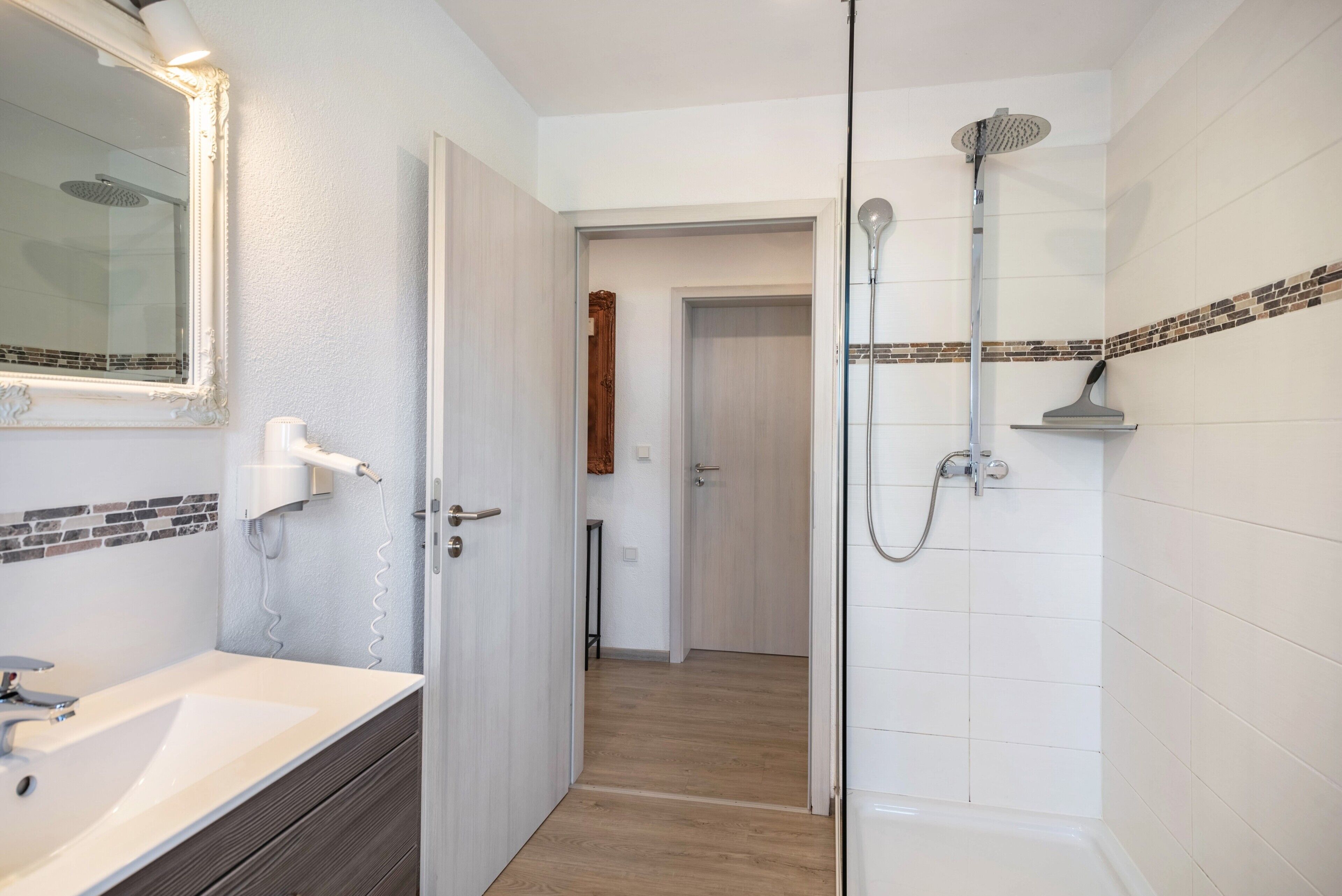 A bathroom with a white sink, a mirror, a shower with a handheld showerhead, and a door leading to another room.