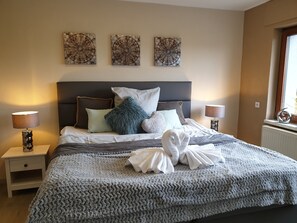 Een net opgemaakt bed met een grijze en witte deken, twee bedlampen en drie kunstwerken aan de muur.