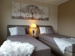 Hotellihuone, jossa on kaksi sänkyä, seinälle ripustettu taideteos, yöpöytä lampun kanssa ja pehmeä valkoinen huopa jokaisen sängyn päällä.