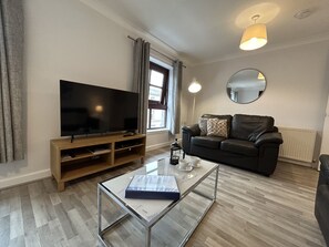 Ein Wohnzimmer mit Flachbildfernseher, Ledersofa, Couchtisch, Spiegel und einem Fenster mit Vorhängen.