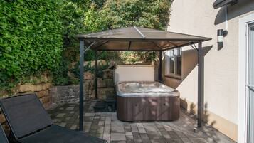Ein Whirlpool unter einer Pergola in einem Garten mit Pflasterterrasse, umgeben von Grünpflanzen.