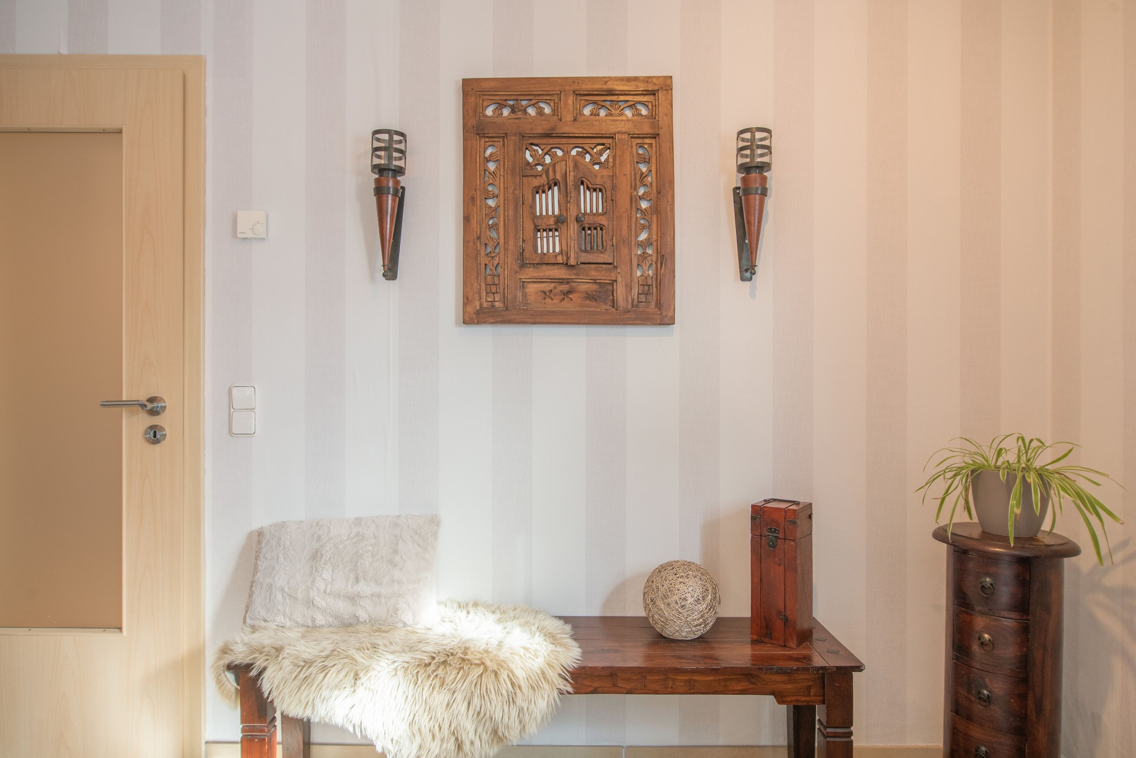 A room with a wooden door, a fluffy chair, a wooden table with a woven ball, a small wooden cabinet, and a potted plant.