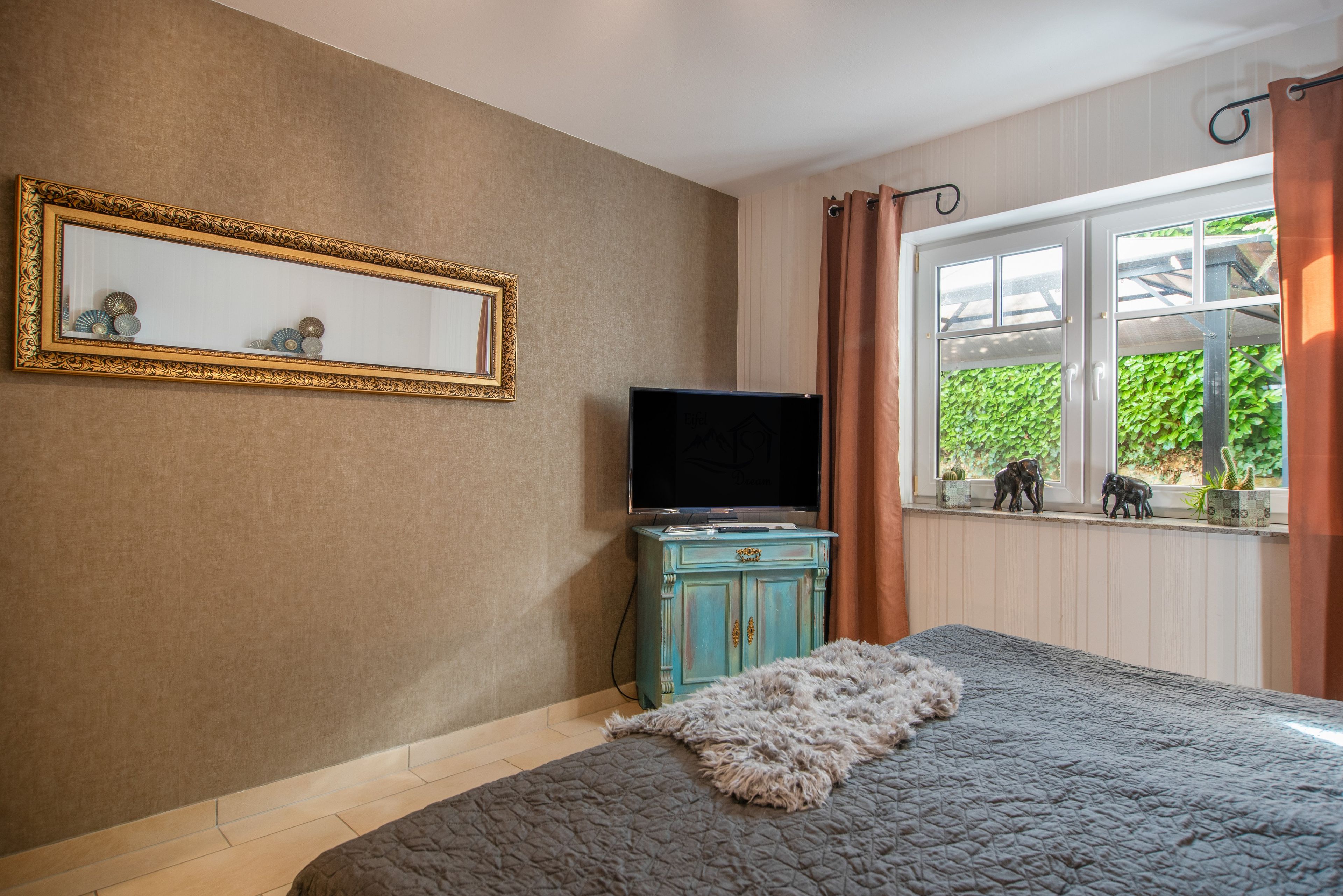 Ein Schlafzimmer mit einem Bett, einem Fernseher auf einer Kommode, einem Spiegel an der Wand und einem Fenster mit Vorhängen.
