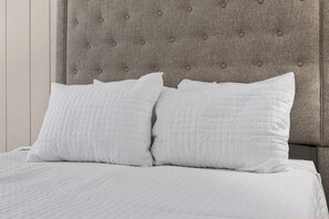 Ein Bett mit weißer Bettwäsche und zwei Kissen, ein grau gepolsterter Kopfteil und eine weiße Wand.