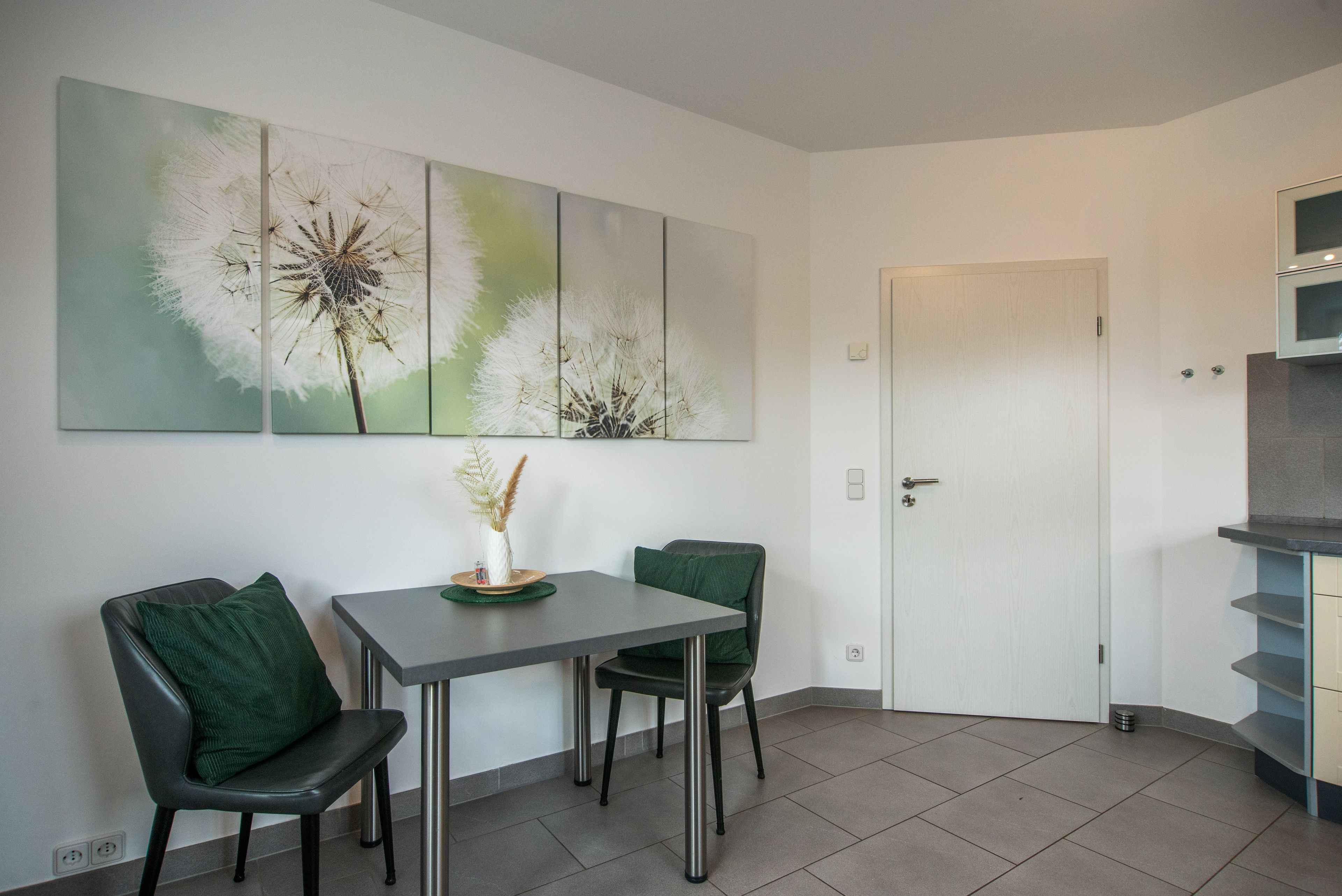 A dining area with a table and chairs, a vase with dried plants, and a wall-mounted artwork of dandelions.