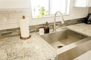 A kitchen with a stainless steel sink, a window with a plant, and a countertop with a bottle and a cup. - Cheerful 2-Bedroom JTree House with Hot Tub and Cowboy Pool (Joshua Tree)