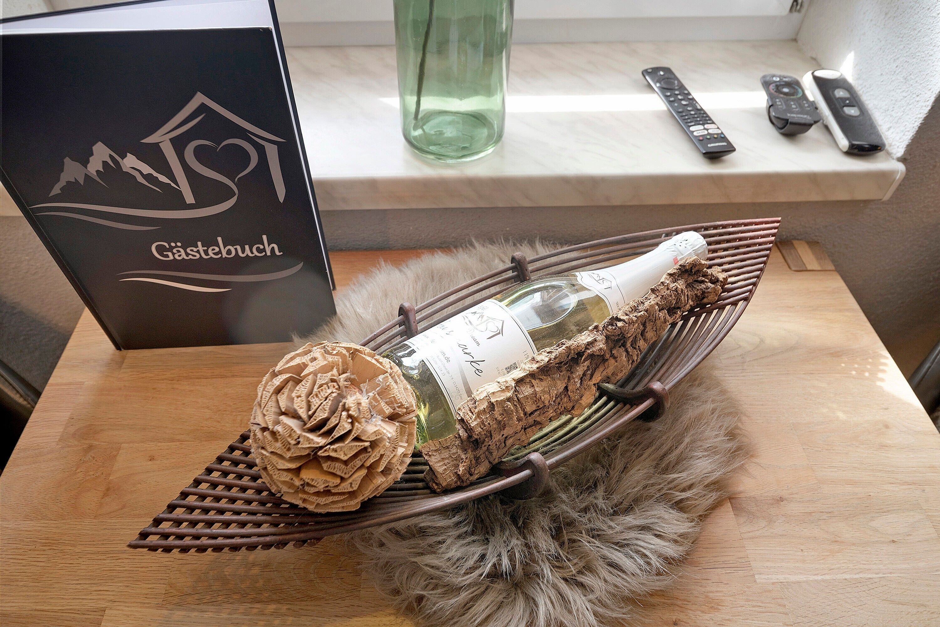 A bottle of wine on a tray with a decorative pine cone and a label, a remote control, and a glass bottle on a shelf.