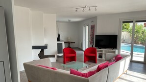 A modern living room with a sofa, red chairs, a glass coffee table, a flat-screen TV, and a view of an outdoor swimming pool. - Villa with pool Between sea and nature (Lattes)