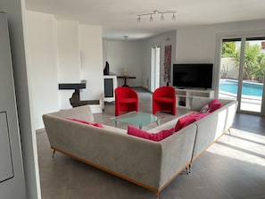 A modern living room with a sofa, red chairs, a glass coffee table, a flat-screen TV, and a view of an outdoor swimming pool. - Villa with pool Between sea and nature (Lattes)