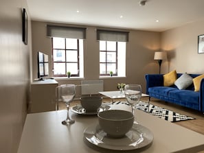 Ein modernes Wohnzimmer mit einem Esstisch für zwei Personen, einem blauen Sofa mit Kissen und großen Fenstern mit Jalousien.