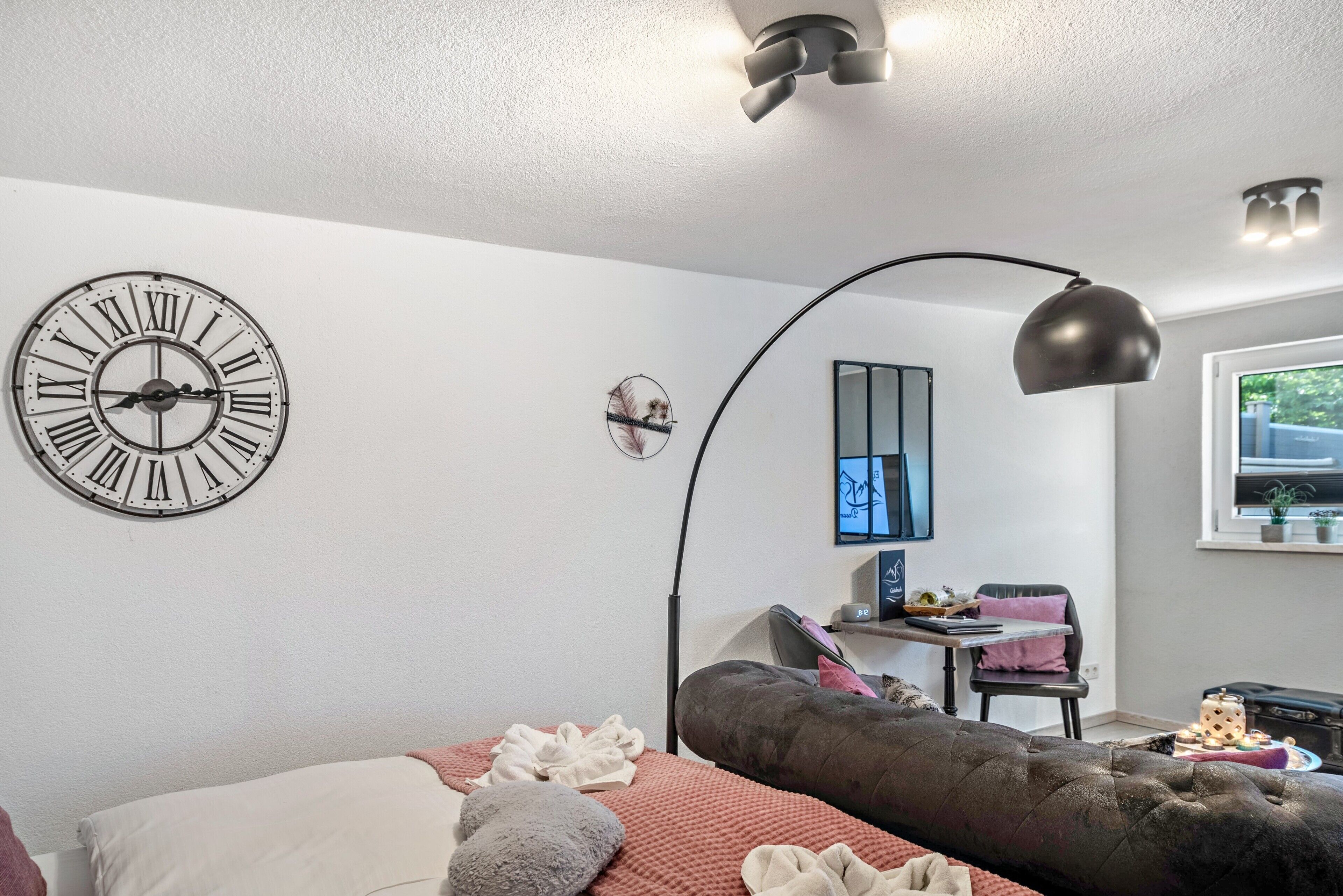Un dormitorio moderno con un reloj de pared grande, una cama con almohadas, un escritorio con una silla y una ventana.