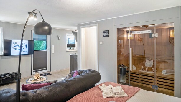 Ett modernt hotellrum med en stor säng, en sittgrupp med en bekväm bänk, en tv och ett spaområde med glasväggar.