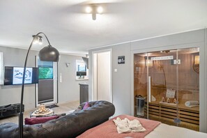 A modern hotel room with a large bed, a seating area with a cushioned bench, a television, and a glass-enclosed spa area. - Eifel Dream Kalkmulde with Private Outdoor Hot Tub and Sauna (Fleringen)