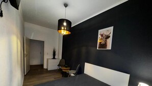Un dormitorio con cama, mesita de noche, lámpara, silla, una pequeña mesa y un cuadro en la pared.