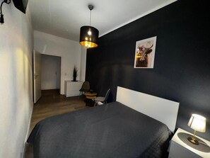 Ein Schlafzimmer mit Bett, Nachttisch, Lampe, Stuhl, kleinem Tisch und einem Bild an der Wand.