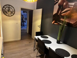 Ein Essbereich mit einem weißen Tisch und schwarzen Stühlen, einer großen Wanduhr und einem Durchgang zu einem anderen Raum.