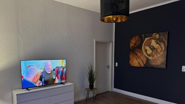 Una sala de estar moderna con una televisión de pantalla plana, un mueble gris, una mesa de centro redonda, una mesita auxiliar con una planta y una pared oscura con un cuadro grande.
