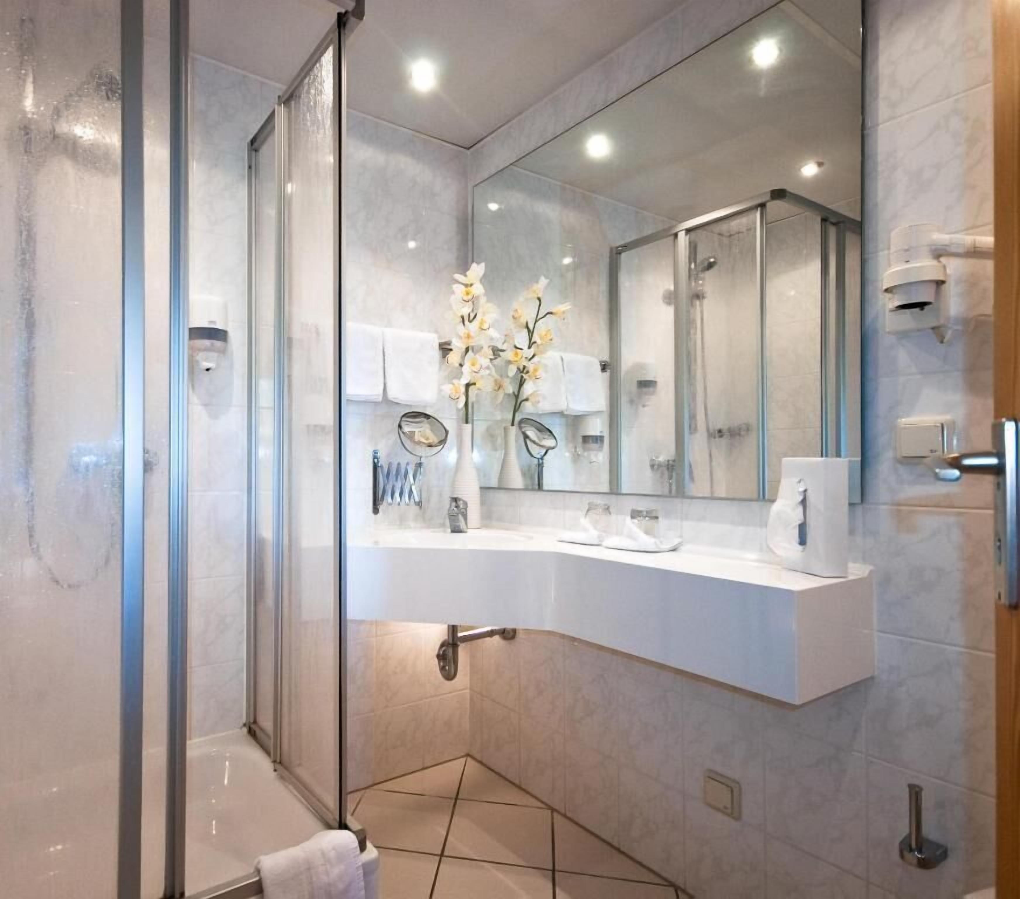 A modern bathroom with a glass shower enclosure, a white vanity with a mirror, and a vase of flowers.