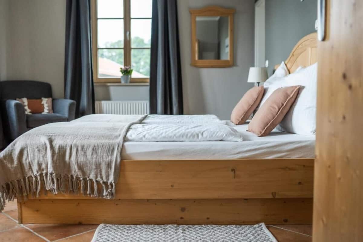 Una cama de madera con cabecera, una mesita de noche, una silla, una ventana con cortinas y un espejo en la pared.