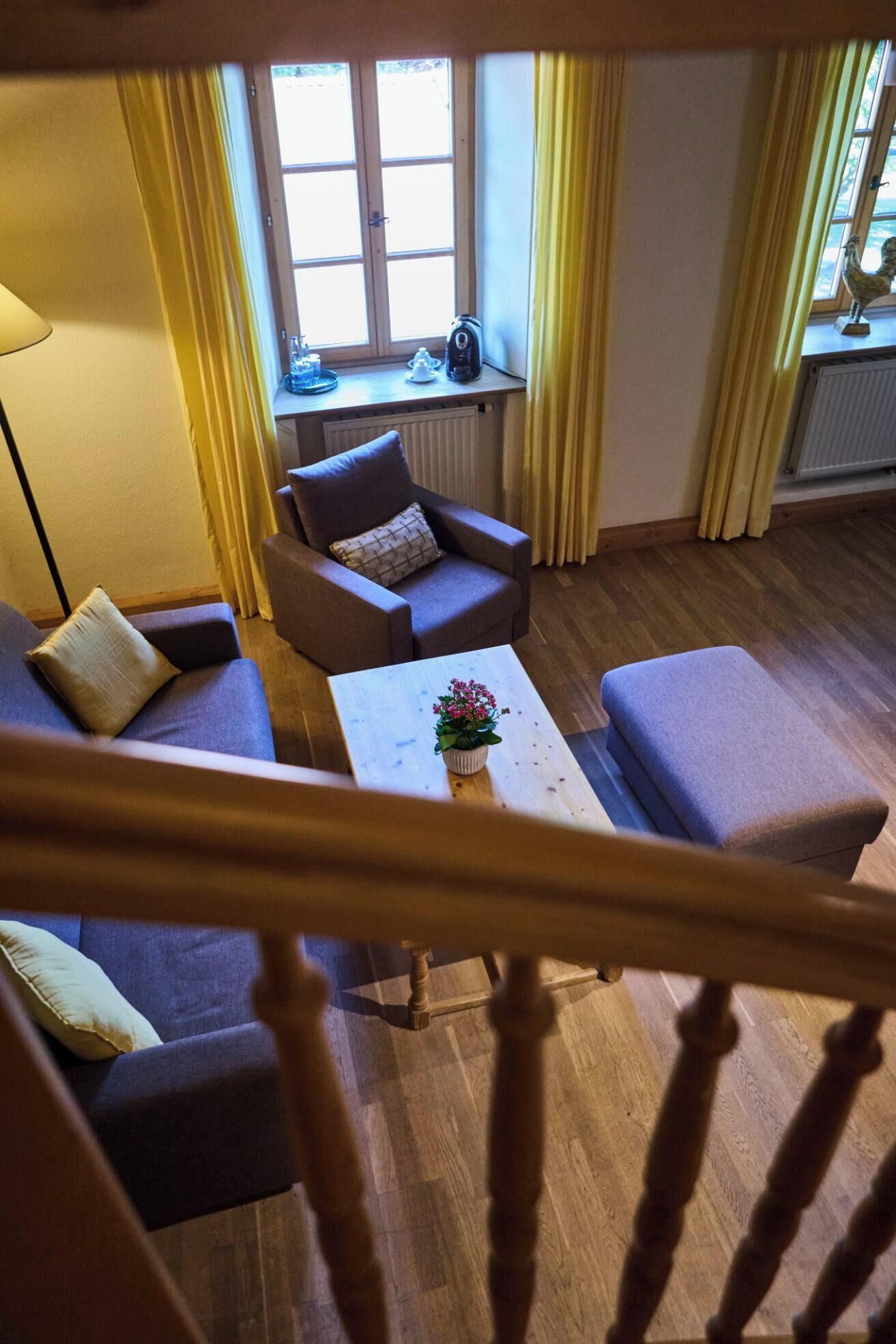 Una habitación con sofá, sillón y reposapiés, una mesa de centro con una flor, y una ventana con cortinas.