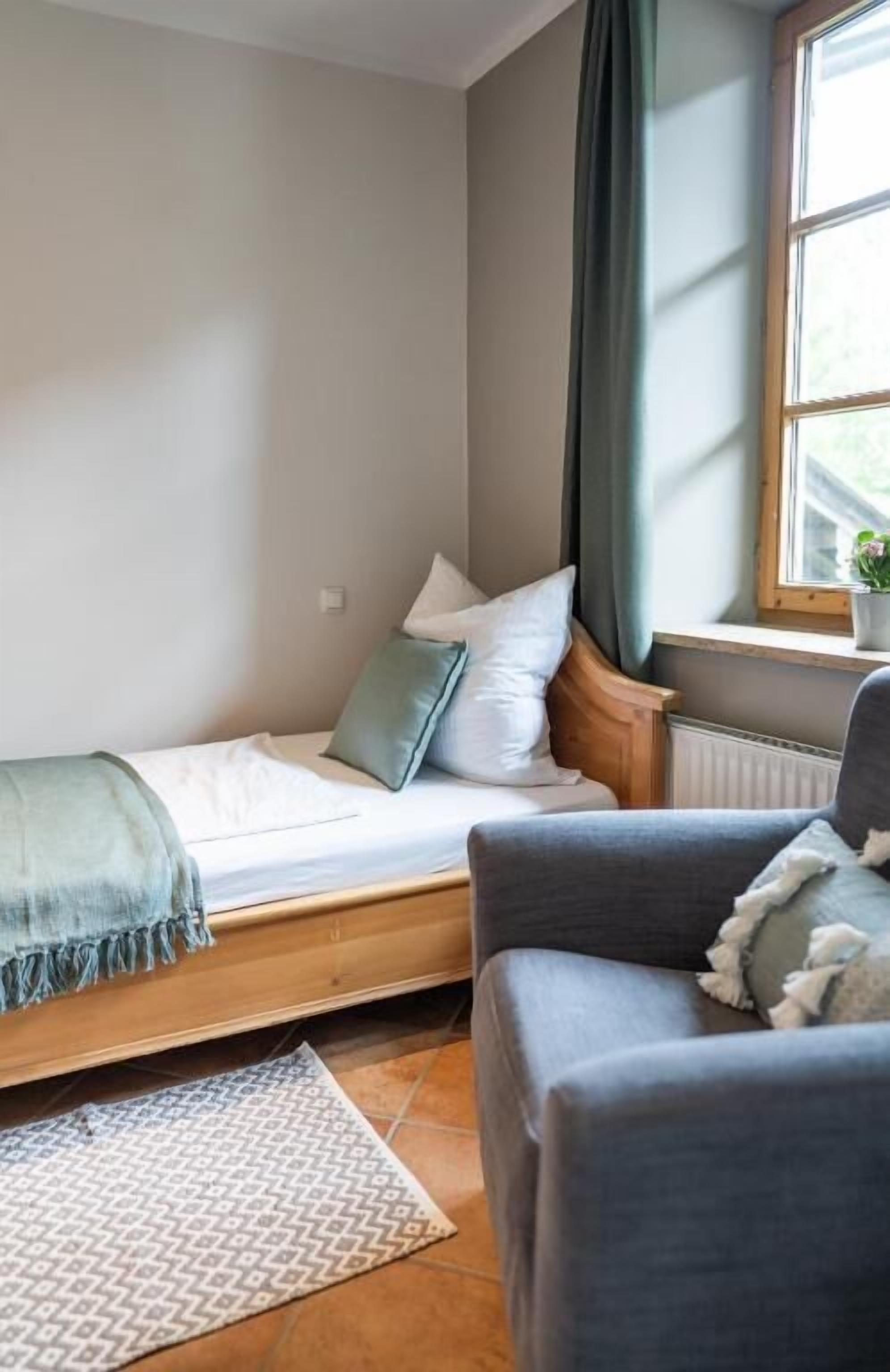 A small, cozy room with a bed, a grey sofa, a window with curtains, and a radiator.