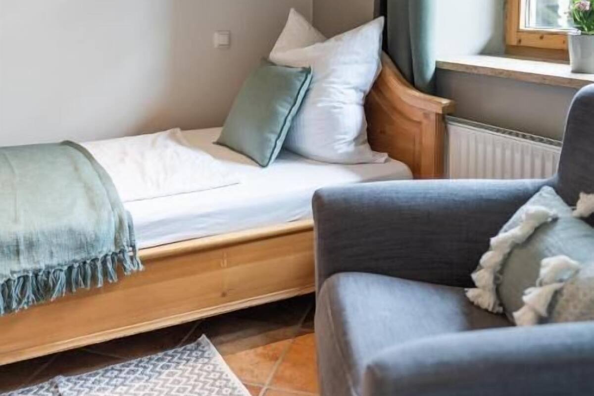 Una habitación pequeña y acogedora con una cama, un sofá gris, una ventana con cortinas y un radiador.