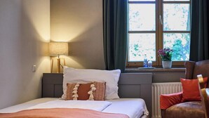 A bedroom with a bed, a chair, a window with curtains, a radiator, and a lamp. - Der Tegernseer Hof (Gmund am Tegernsee)
