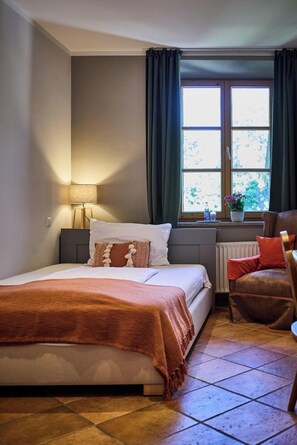 Quarto com cama, cadeira, janela com cortinas, radiador e luminária.