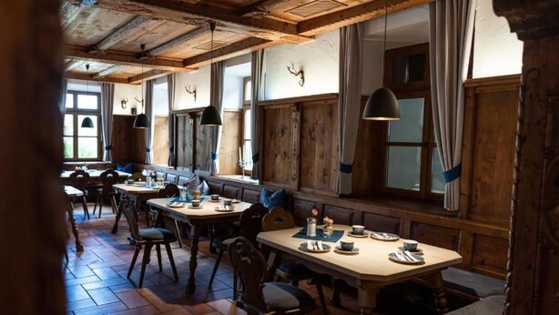Een rustieke eetruimte met houten tafels en banken, grote ramen met gordijnen en een houten plafond.