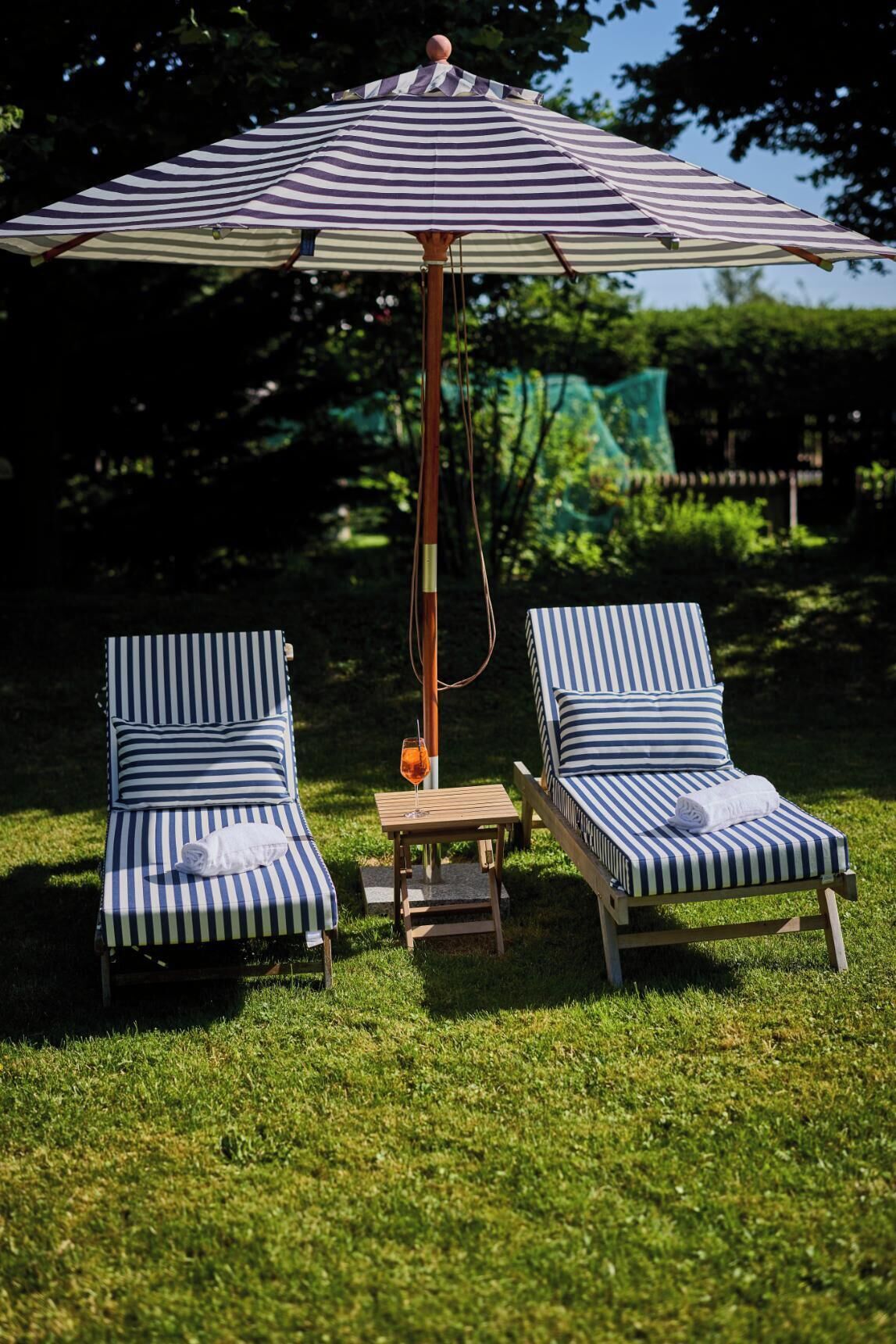 Two striped lounge chairs with a small table and a striped umbrella in a grassy outdoor area.