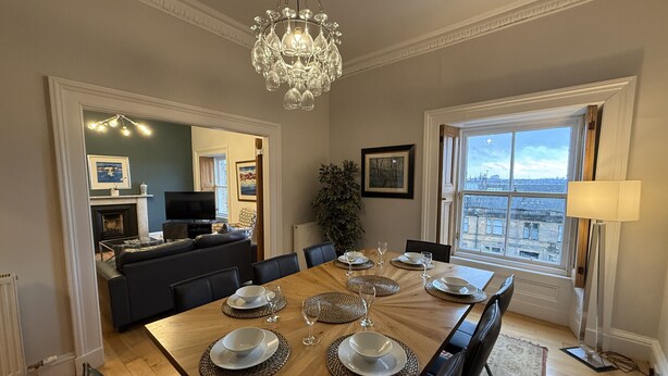 A dining area with a wooden table set for six, surrounded by black chairs, a chandelier, and a view through a large window. - Crown Circus Three Bedroom Apartment (Hillhead)