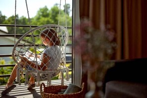 Une personne assise dans un fauteuil en osier près d’une fenêtre, avec vue sur la verdure à l’extérieur.