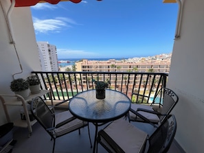Ein Balkon mit rundem Tisch und Stühlen, Blumentöpfen und Blick auf das Meer und die Gebäude.