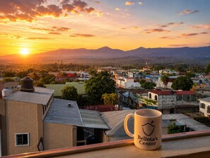Une tasse ornée de l’inscription « Bellota’s Center », posée sur un balcon donnant sur un paysage urbain avec des bâtiments et des montagnes, au coucher du soleil.